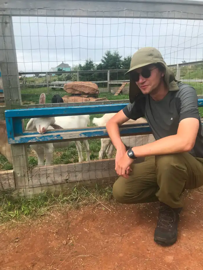 L'auteur est accroupi devant une clôture grillagée derrière laquelle se trouve une chèvre.